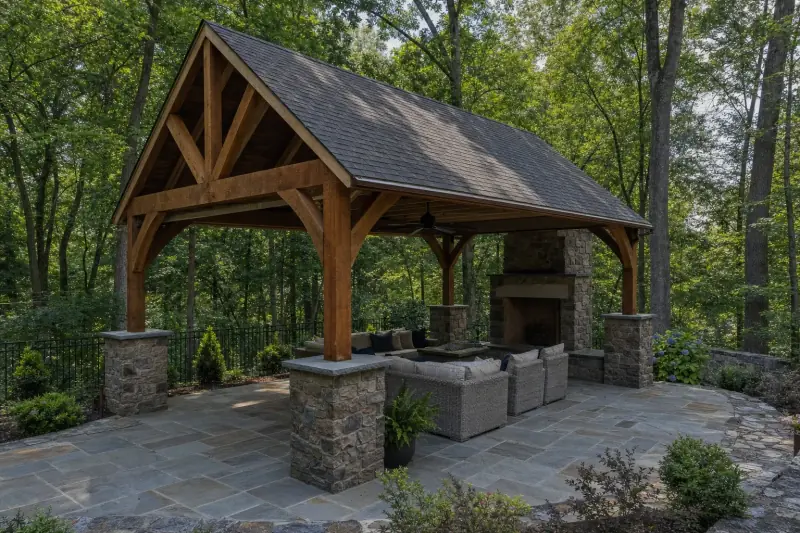 Timber pavilion with a shingled roof and a stone fireplace over a patio
